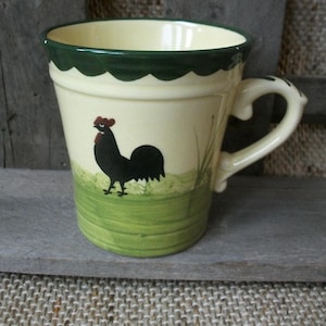 Könnte beinhalten: Cremefarbene Keramiktasse mit einer schwarzen Hahn-Illustration auf einer grünen Grasbasis. Die Tasse hat einen gezackten dunkelgrünen Rand und einen gebogenen Griff. Die Tasse ist etwa 10 cm hoch.