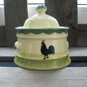 Könnte beinhalten: Gelbe Keramikdose mit Deckel und grünen Verzierungen. Auf der Vorderseite ist ein schwarzer Hahn abgebildet. Die Dose hat einen grünen, gewellten Rand und einen kleinen Knopf auf dem Deckel. Geeignet für die Küche, ist die Dose ein dekoratives Element.