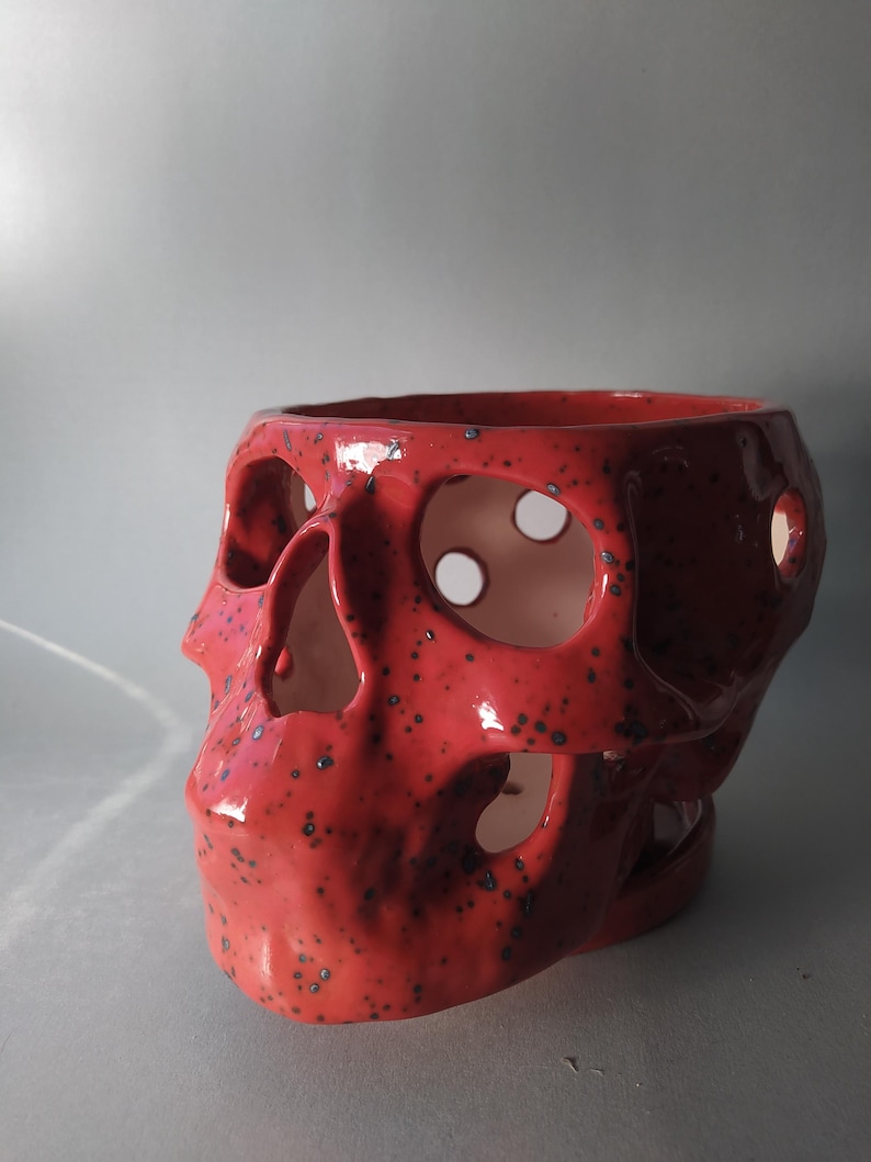 Puede incluir: Un recipiente de cer&aacute;mica con forma de cr&aacute;neo, acabado en esmalte rojo brillante con motas azul oscuro. El objeto presenta aberturas circulares recortadas en los ojos, la nariz y los laterales, dejando ver una superficie interior blanca y lisa.