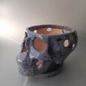 Puede incluir: Una maceta de cer&aacute;mica con forma de cr&aacute;neo, con un esmalte de color morado claro y motas de color verde azulado oscuro. El objeto presenta recortes circulares en todo el cr&aacute;neo y las cuencas de los ojos, revelando una superficie interior lisa.