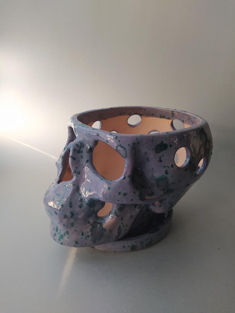 Puede incluir: Una maceta de cer&aacute;mica con forma de cr&aacute;neo, con un esmalte de color morado claro y motas de color verde azulado oscuro. El objeto presenta recortes circulares en todo el cr&aacute;neo y las cuencas de los ojos, revelando una superficie interior lisa.