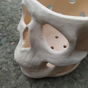 May include: A cream-colored ceramic skull-shaped planter. The planter has large eye and nose openings, and several small circular drainage holes. The skull's surface has a textured, matte finish, and the overall design is a unique decorative item.
