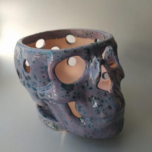 Puede incluir: Un recipiente de cer&aacute;mica con forma de calavera que presenta un esmalte de color morado claro con motas de color verde azulado oscuro. El objeto tiene recortes circulares en el cr&aacute;neo y la zona facial, revelando una superficie interior clara.