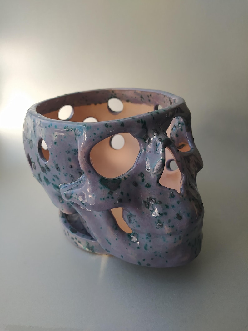 Puede incluir: Un recipiente de cer&aacute;mica con forma de calavera que presenta un esmalte de color morado claro con motas de color verde azulado oscuro. El objeto tiene recortes circulares en el cr&aacute;neo y la zona facial, revelando una superficie interior clara.