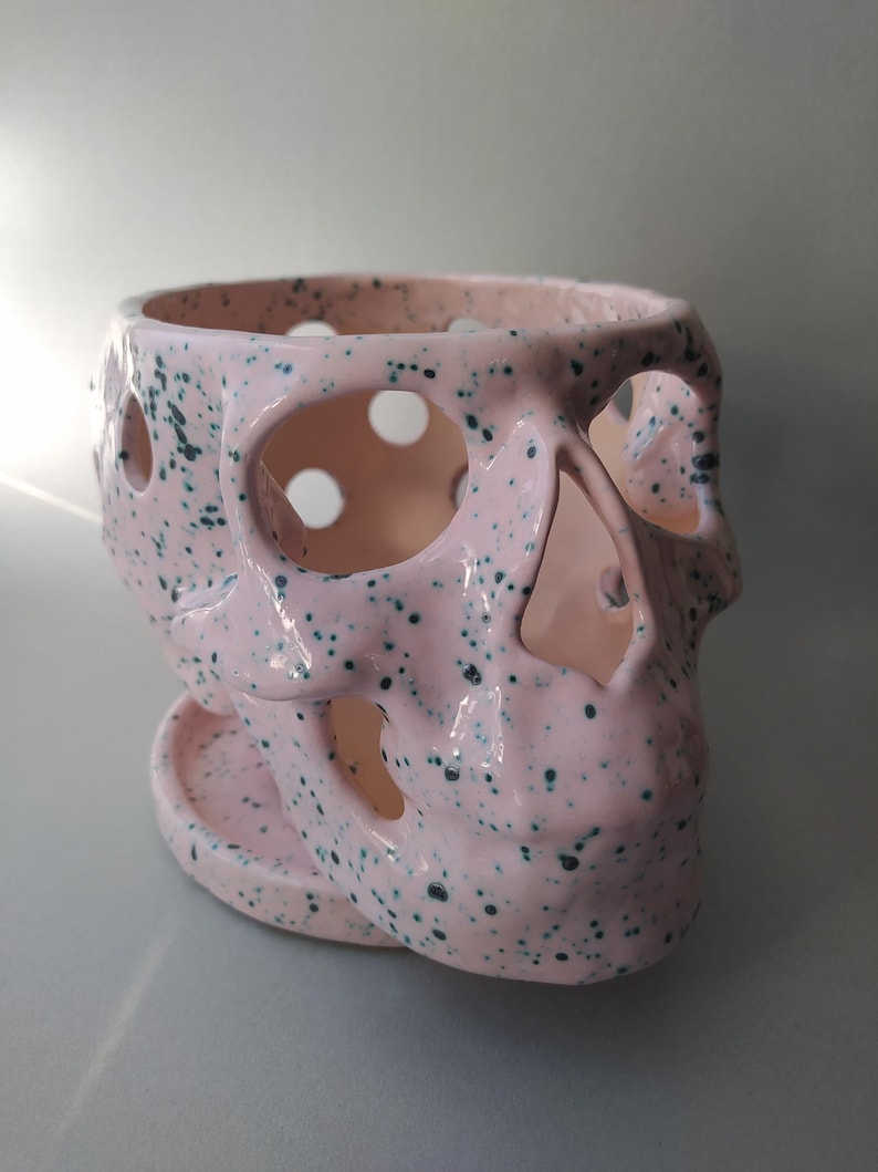 Puede incluir: Una maceta de cer&aacute;mica con forma de cr&aacute;neo, esmalte rosa claro brillante y motas de color verde oscuro. La maceta presenta cuencas oculares, cavidad nasal y aberturas circulares en el cr&aacute;neo, y descansa sobre un plato rosa a juego.