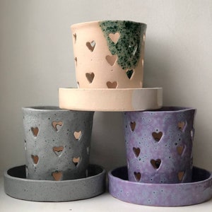 May include: Three ceramic planters with heart-shaped cutouts. The planters are in shades of gray, purple, and pink with green speckles. Each planter has a matching saucer.