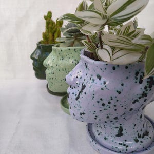 Könnte beinhalten: Drei Keramik-Gesichtspflanzgefäße in einer Reihe, jedes mit einer anderen Pflanze. Die Pflanzgefäße sind in Grün- und Lavendeltönen gehalten und mit dunkelgrünen Punkten besprenkelt. Zu den Pflanzen gehören ein Kaktus und eine Pflanze mit weiß-grün gestreiften Blättern.