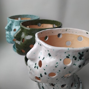 Può includere: Tre vasi in ceramica a forma di volto. Il vaso anteriore è bianco con macchie verdi. Il secondo vaso è verde scuro e il terzo azzurro. Ogni vaso ha fori circolari e una base rotonda. I vasi sono per le piante.