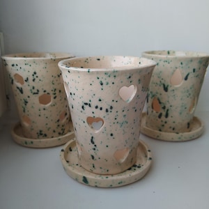 May include: Three ceramic flower pots with matching saucers. The pots are speckled with teal and have heart-shaped cutouts. The pots are a light beige color.