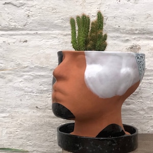 Op de afbeelding: Een keramische bloempot in de vorm van een menselijk hoofd, met een terracotta gezicht en een wit en blauw gespikkeld ontwerp. Het bevat een kleine groene cactus. De basis is zwart en gespikkeld. De pot staat tegen een witte bakstenen muur.