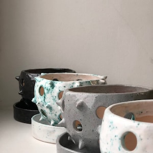 May include: Four ceramic planters with a spiky, textured exterior and drainage holes. The planters are in various colours, including black, white with green speckles, grey, and white with blue speckles. Each planter has a matching saucer.
