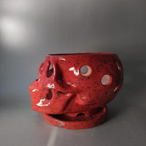 Puede incluir: Una maceta de cer&aacute;mica de color rojo brillante con forma de cr&aacute;neo humano, acabado brillante y motas oscuras. El cr&aacute;neo presenta m&uacute;ltiples aberturas circulares en su superficie y descansa sobre una base de platillo de cer&aacute;mica roja a juego.