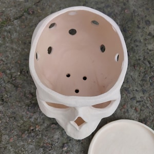 May include: A cream-colored skull-shaped planter with multiple drainage holes in the top. The planter has a matching saucer. The skull's eye sockets and nose cavity are visible. The planter is made of ceramic.