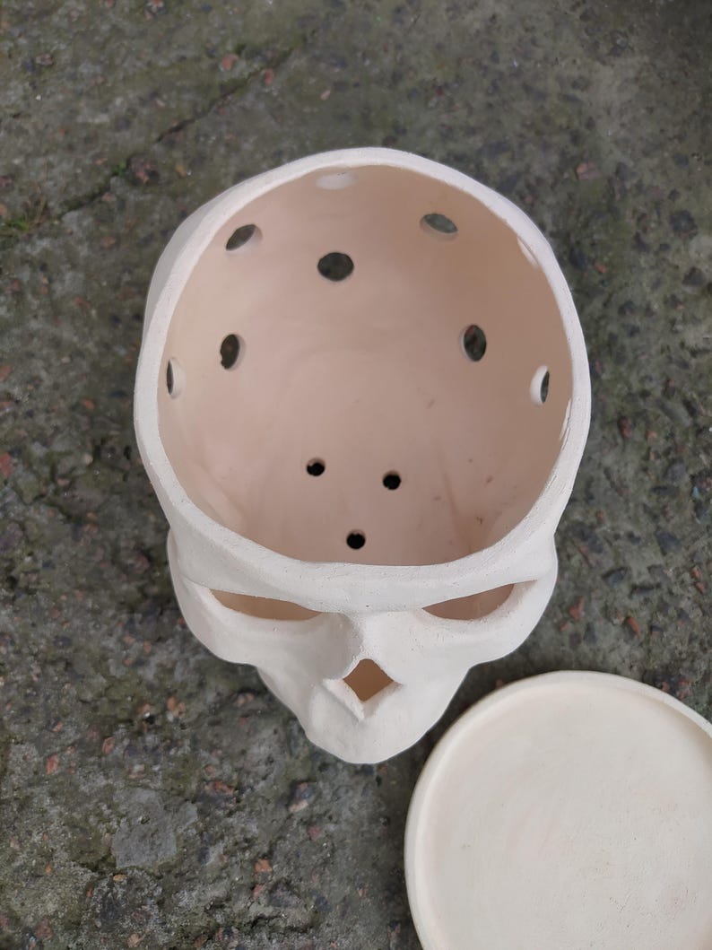 May include: A cream-colored skull-shaped planter with multiple drainage holes in the top. The planter has a matching saucer. The skull's eye sockets and nose cavity are visible. The planter is made of ceramic.