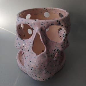 Puede incluir: Una maceta de cer&aacute;mica de color rosa claro con forma de calavera y un patr&oacute;n de motas de color verde oscuro. El objeto tiene grandes aberturas circulares para los ojos, la nariz y el cr&aacute;neo, e incluye una base circular a juego para el drenaje.