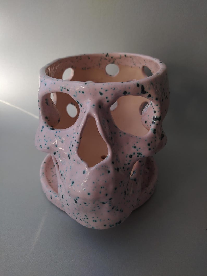 Puede incluir: Una maceta de cer&aacute;mica de color rosa claro con forma de calavera y un patr&oacute;n de motas de color verde oscuro. El objeto tiene grandes aberturas circulares para los ojos, la nariz y el cr&aacute;neo, e incluye una base circular a juego para el drenaje.