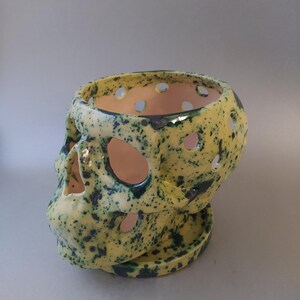 Puede incluir: Una maceta de cer&aacute;mica con forma de calavera que presenta un esmalte amarillo brillante con motas azul oscuro. El objeto incluye recortes circulares en el cr&aacute;neo y las cuencas de los ojos, y descansa sobre una base de plato circular a juego.