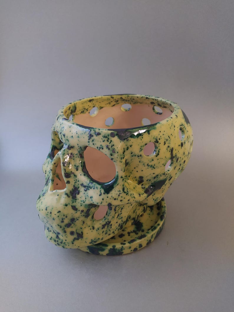 Puede incluir: Una maceta de cer&aacute;mica con forma de calavera que presenta un esmalte amarillo brillante con motas azul oscuro. El objeto incluye recortes circulares en el cr&aacute;neo y las cuencas de los ojos, y descansa sobre una base de plato circular a juego.