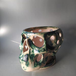 Puede incluir: Un recipiente de cer&aacute;mica con forma de cr&aacute;neo que presenta un esmalte base de color blanco roto brillante con acentos moteados en verde oscuro y azul. El objeto tiene recortes circulares en todo el cr&aacute;neo y la zona facial, creando una estructura.