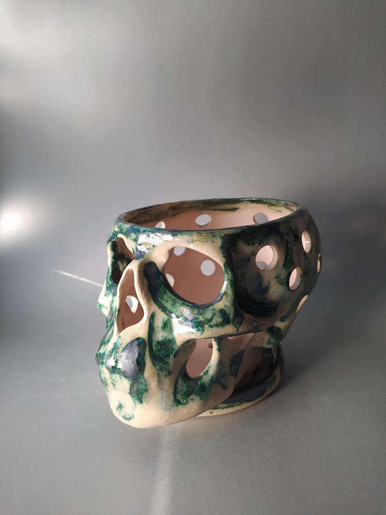 Puede incluir: Un recipiente de cer&aacute;mica con forma de cr&aacute;neo que presenta un esmalte base de color blanco roto brillante con acentos moteados en verde oscuro y azul. El objeto tiene recortes circulares en todo el cr&aacute;neo y la zona facial, creando una estructura.