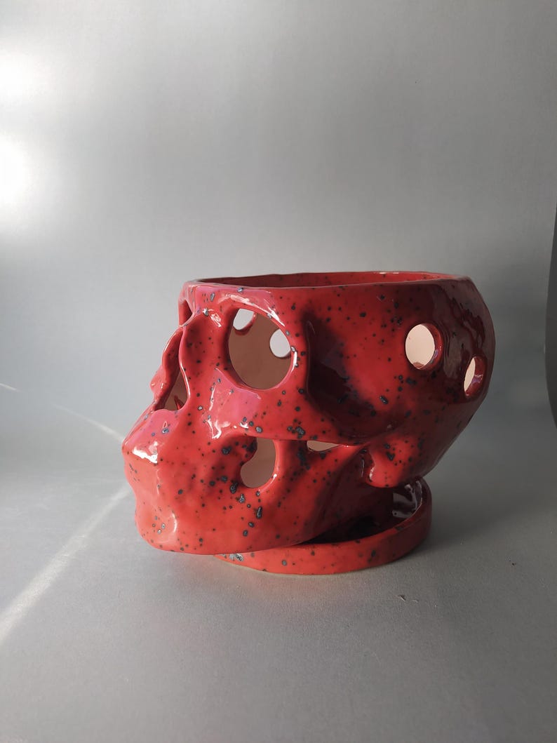 Puede incluir: Una maceta de cer&aacute;mica roja con forma de calavera, acabado brillante y motas azul oscuro. La calavera presenta cuencas oculares y cavidades nasales recortadas, y descansa sobre una base de platillo circular de cer&aacute;mica roja a juego.