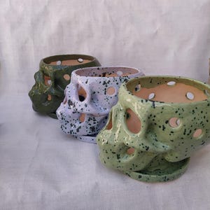 May include: Three ceramic skull planters in shades of green and lavender, speckled with darker spots. Each skull has eye and nose openings, and a circular opening at the top. The planters are arranged in a row, showcasing their unique design.