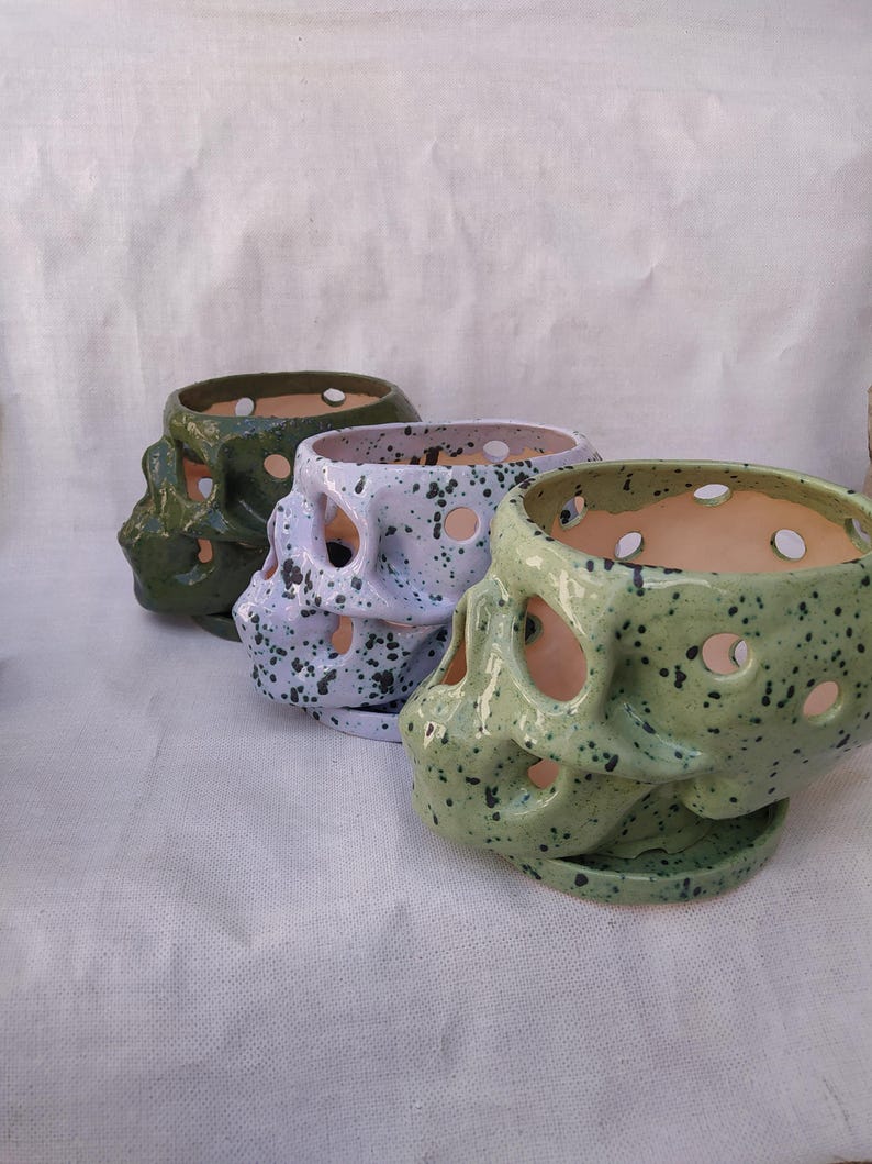 Puede incluir: Tres macetas de cer&aacute;mica con forma de calavera dispuestas en fila. La maceta delantera es de color verde claro con motas negras, la del medio es de color violeta claro con motas negras y la trasera es de color verde oliva oscuro brillante.