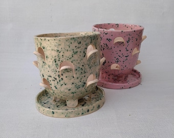 May include: Two ceramic planters with matching saucers. One planter is cream-coloured with dark green speckles, and the other is pink with dark green speckles. Both feature multiple arched cutouts along the sides and a smooth, glossy finish.