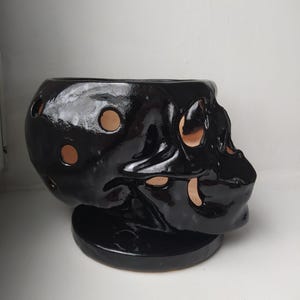 Puede incluir: Un recipiente de cer&aacute;mica negra brillante con forma de cr&aacute;neo que presenta m&uacute;ltiples recortes circulares en su superficie. El interior del cr&aacute;neo no est&aacute; esmaltado, revelando un acabado de arcilla color terracota natural visible a trav&eacute;s de las aberturas.
