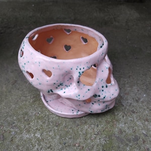 Puede incluir: Una maceta de cerámica rosa con forma de calavera, con recortes en forma de corazón y motas verdes. La maceta tiene una base redonda y una gran abertura. Las cuencas de los ojos y la nariz de la calavera están abiertas.