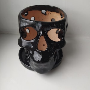 Puede incluir: Una maceta de cer&aacute;mica negra con forma de calavera, acabado brillante y plato circular a juego. El interior de la calavera es de arcilla marr&oacute;n claro sin esmaltar, con cuencas oculares, cavidad nasal y orificios circulares en el borde superior.