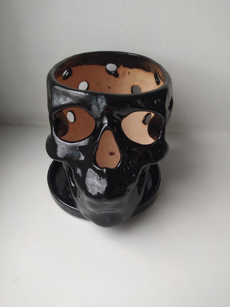 Puede incluir: Una maceta de cer&aacute;mica negra con forma de calavera, acabado brillante y plato circular a juego. El interior de la calavera es de arcilla marr&oacute;n claro sin esmaltar, con cuencas oculares, cavidad nasal y orificios circulares en el borde superior.