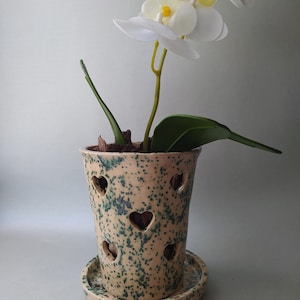 May include: A ceramic orchid pot with a speckled beige and green glaze featuring heart-shaped cutouts throughout the body. The pot sits on a matching circular saucer and contains a white artificial orchid plant with green leaves and brown bark.