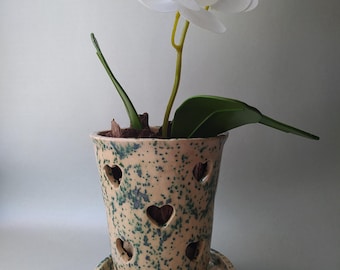 May include: A ceramic orchid pot with a speckled beige and green glaze featuring heart-shaped cutouts throughout the body. The pot sits on a matching circular saucer and contains a white artificial orchid plant with green leaves and brown bark.