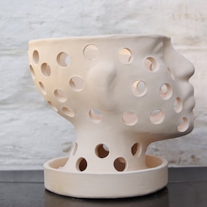 May include: A cream-colored ceramic planter shaped like a human head with numerous circular cutouts. The planter has a detailed profile and sits on a circular base. The design is modern and artistic, suitable for indoor decor.