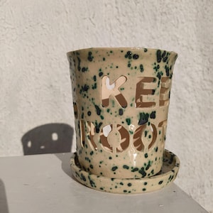 Könnte beinhalten: Beigefarbener Keramik-Pflanztopf mit passender Untertasse, verziert mit dunkelgrünen Sprenkeln. Der Topf hat ausgeschnittene Buchstaben mit der Aufschrift "KEEP ROOTING". Der Pflanztopf steht auf einer weißen Oberfläche vor einer strukturierten Wand.