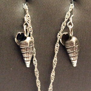 Könnte beinhalten: Silberfarbenes Schmuckset mit Halskette und Ohrringen. Die Ohrringe und der Anhänger haben ein Muscheldesign. Die Kette ist im gedrehten Seil-Stil. Der Schmuck wird vor dunklem Hintergrund präsentiert.