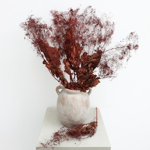 May include: A still life composition showing a weathered, off-white ceramic vase with two handles, filled with dried, reddish-brown foliage. The vase is placed on a square, light-coloured pedestal, with fallen leaves around its base. The background is plain white.
