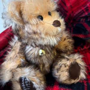 May include: A vintage teddy bear with brown and tan fur, black eyes, and a small bell on its collar. The bear is sitting on a red and black plaid blanket. The bear's paw has the text " Joanne 1992".