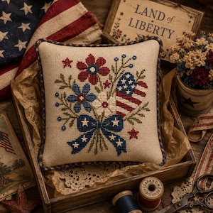May include: A square decorative pillow with a beige linen-like fabric featuring a cross-stitched bouquet of red, white, and blue flowers, a heart-shaped American flag, and a blue bow with white stars, finished with a dark blue and brown braided trim.