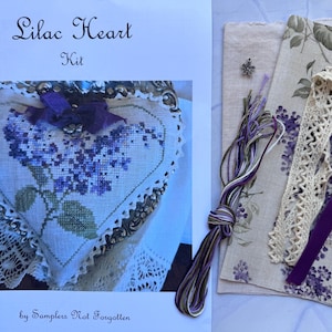 May include: A cross-stitch kit titled "Lilac Heart" featuring a heart-shaped design with a lilac flower pattern. The kit includes fabric, thread, lace trim, and a purple ribbon. The text "by Samplers Not Forgotten" is visible.