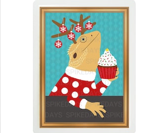 Peut inclure: Un lézard barbu portant un pull rouge à pois blancs avec un bonnet de Père Noël rouge et des bois de renne. Le lézard tient un cupcake rouge et blanc avec des pépites vertes et une étoile au sommet. L'arrière-plan est bleu turquoise avec un motif blanc.