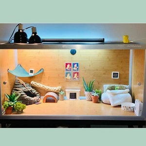以下が含まれることがあります： 木製の背景と、小さなサボテン、葉の植物、小さな木など、さまざまな植物のあるガラスのテラリウム。トカゲ用の白いハンモック、小さな岩の形成、小さな木の橋があります。テラリウムは、2つの黒いヒートランプで照らされています。