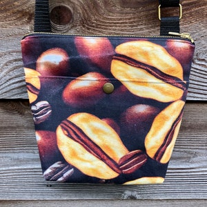 Op de afbeelding: Een klein zwart canvas tasje met een ritssluiting en een gouden knoop. De tas heeft een bruine schouderband en een print van koffiebonen.