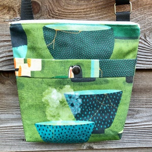 May include: A green fabric crossbody bag with a zipper closure and a flap with a button. The bag features a repeating pattern of blue and green bowls with white polka dots and gold accents.