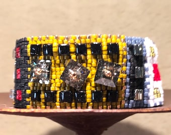 May include: A wide beaded bracelet with a geometric pattern. The bracelet features yellow, black, red, and grey beads, with three square, clear, faceted embellishments. The bracelet is approximately 3.8 cm wide.