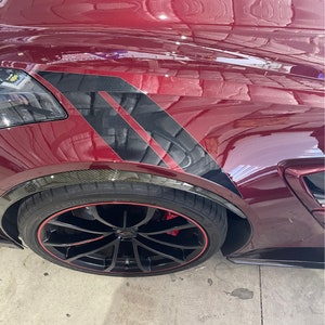 Puede incluir: Primer plano de un coche deportivo rojo con rayas de carreras negras en el guardabarros delantero. El coche tiene ruedas negras con un borde rojo y un acento de fibra de carbono en el guardabarros.