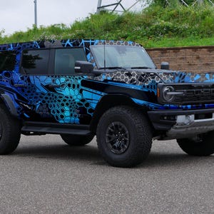 Pode incluir: Um Ford Bronco azul e preto com um envolt&oacute;rio de camuflagem. O ve&iacute;culo tem pneus grandes e uma grelha preta.