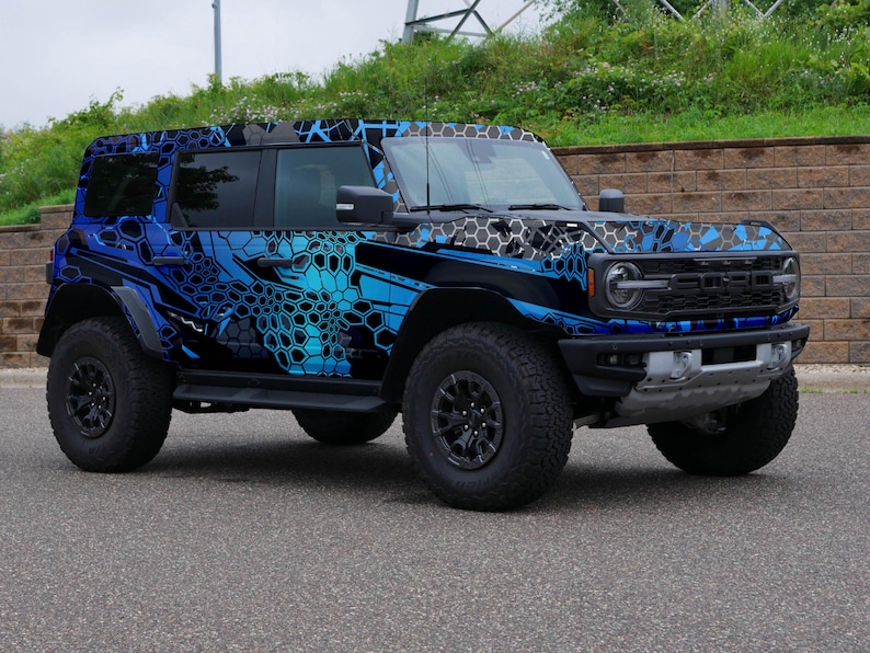 Pode incluir: Um Ford Bronco azul e preto com um envolt&oacute;rio de camuflagem. O ve&iacute;culo tem pneus grandes e uma grelha preta.