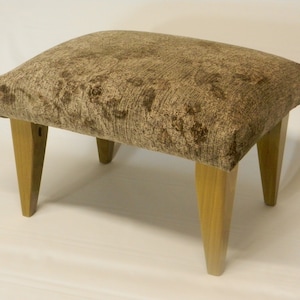 May include: A brown upholstered footstool with four wooden legs. The fabric has a subtle pattern.